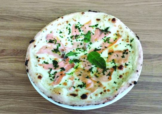Pizza blanche garnie de saumon, crème fraîche et herbes, sur une assiette blanche.