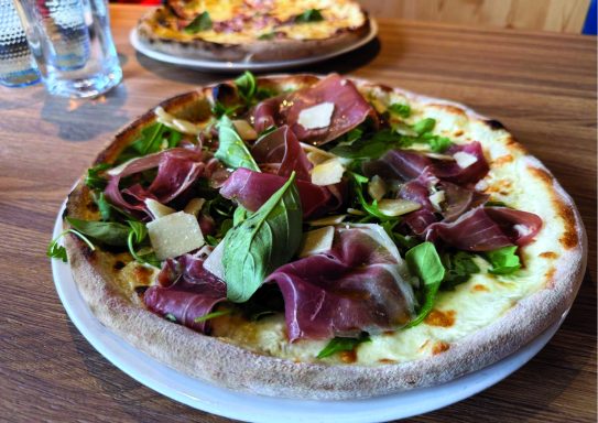 Pizza garnie de prosciutto, roquette et copeaux de fromage sur une assiette en bois.